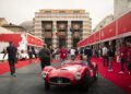 A vintage red Maserati race car with two drivers is parked on a red carpeted street during the 1000 Miglia, surrounded by people and event banners in front of a large building with Italian and EU flags.