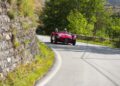 A red vintage Maserati race car with two people in helmets drives on a winding road surrounded by greenery and a stone wall, capturing the spirit of the 1000 Miglia.