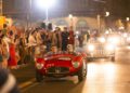 A vintage red Maserati race car with two drivers moves through a lit street at night, followed by another classic car, as crowds watch from the sidewalk during the legendary 1000 Miglia.