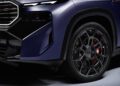 Close-up view of the front left side of a dark blue 2026 BMW XM, showcasing its headlight, bold grille, and a black alloy wheel with striking red brake calipers.