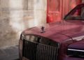 A maroon Rolls-Royce Spectre with a European license plate is parked by a stone wall and large red doors, exuding the elegance of Paris Fashion Week.