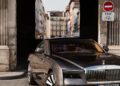 A gray Rolls-Royce Spectre is parked in front of a historic stone archway, with buildings and a no entry sign visible in the background, capturing timeless elegance reminiscent of Paris Fashion Week.