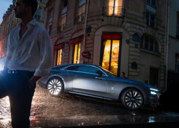 A man in a white shirt stands in the foreground on a wet street, while a gray Cullinan luxury car is parked in front of a building on a rainy evening, evoking the elegance of Paris Fashion Week.