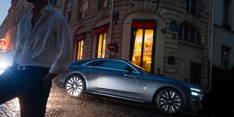 A man in a white shirt stands in the foreground on a wet street, while a gray Cullinan luxury car is parked in front of a building on a rainy evening, evoking the elegance of Paris Fashion Week.