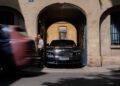 A man stands beside a parked black Rolls-Royce Spectre in an arched building entrance as a red vehicle passes by on the street, capturing the essence of Paris Fashion Week style.