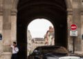 A man in formal attire stands by a stone archway while a Rolls-Royce with a European license plate is parked nearby, evoking the elegance of Paris Fashion Week.