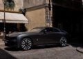 A black Rolls-Royce Spectre coupe is parked on a cobblestone street beside a café with outdoor seating, capturing the elegance and style of Paris Fashion Week in a European city.