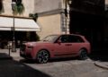 A pink Rolls-Royce Spectre SUV is parked on a cobblestone street in front of a café with outdoor seating and a beige awning, radiating Paris Fashion Week elegance.