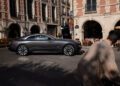 A gray Rolls-Royce Spectre coupe is parked on a city street near outdoor café seating, capturing the chic atmosphere reminiscent of Paris Fashion Week as a person strolls by in the foreground.