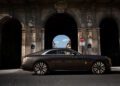 A Rolls-Royce Spectre luxury coupe parked in front of a historic stone archway under daylight, with a blue sky and buildings visible in the background—an elegant scene fit for Paris Fashion Week.