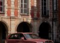 A maroon Rolls-Royce Spectre SUV is parked on a street in front of a historic building with large windows and arched doorways, capturing the elegance of Paris Fashion Week.