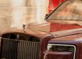 A maroon Rolls-Royce Spectre with a visible license plate is parked near a beige wall and a red door.