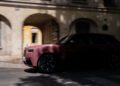 A maroon Cullinan luxury SUV drives past a beige building with arched windows and shadows cast on the facade, exuding an elegance fit for Paris Fashion Week.