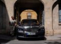 A man in sunglasses leans against a building archway next to a parked black Rolls-Royce Spectre with a visible license plate, in an urban setting.