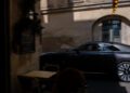 A black Rolls-Royce glides past a table and chairs in a shaded outdoor area near a beige building with arched windows, capturing the elegance of Paris Fashion Week.