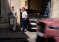 Two men stand in front of a parked Rolls-Royce under an archway, as a pink car passes by on the street in bright sunlight—a scene reminiscent of Paris Fashion Week.