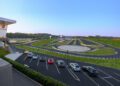 A modern driving course at One Porsche Drive in Atlanta features multiple test tracks and a parking lot in the foreground containing several parked Porsche cars under clear blue skies.