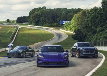 Three Porsche vehicles, including a purple Taycan, are driving on a winding racetrack surrounded by grass and trees.