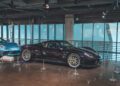 A dark purple Porsche 918 Spyder is displayed indoors next to a blue Porsche sports car, separated by ropes in a modern showroom at One Porsche Drive in Atlanta.