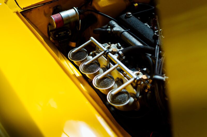 Close-up of a yellow Alfa Romeo Giulia GTV’s engine bay, as featured on Petrolicious Film Friday, showcasing detailed carburetor components and intricate metal piping.