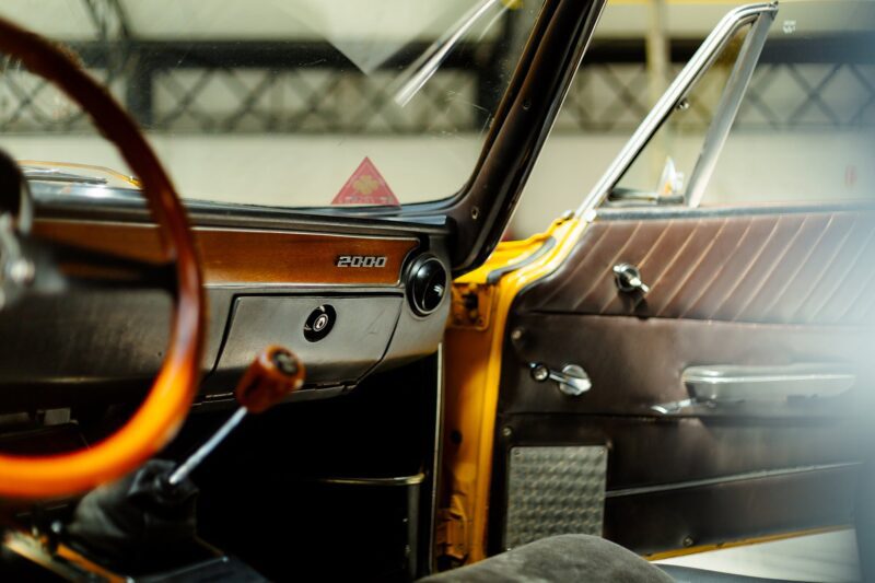 Close-up of an Alfa Romeo Giulia GTV interior showing a wooden steering wheel, gear shift, brown door panel, and "2000" badge on the dashboard—perfect for a Film Friday feature on Petrolicious.