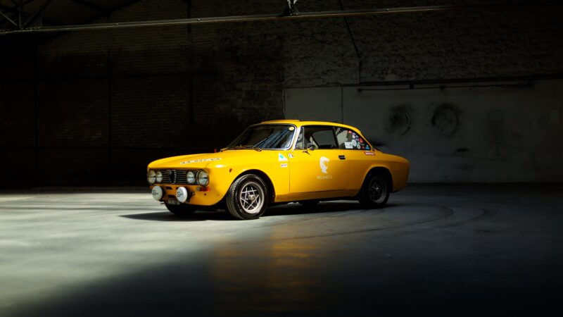 A vintage yellow Alfa Romeo Giulia GTV is parked in a large, dimly lit industrial space, with light highlighting the vehicle—a true Petrolicious scene.