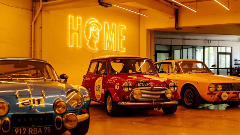 Three vintage cars—a blue Alpine, a red Mini Cooper, and a yellow Alfa Romeo Giulia GTV—parked indoors under a neon "HOME" sign on a yellow wall. Perfect for Film Friday or any Petrolicious enthusiast.