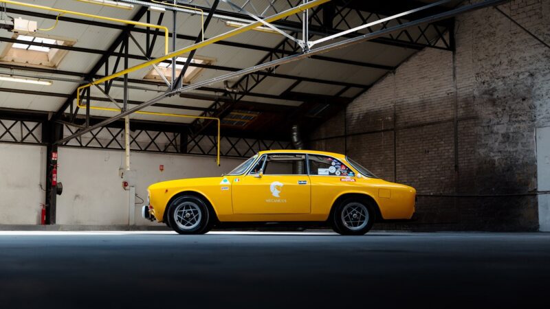A yellow vintage Alfa Romeo Giulia GTV with decals is parked inside an industrial warehouse with concrete walls and skylights, perfect for a Petrolicious Film Friday feature.