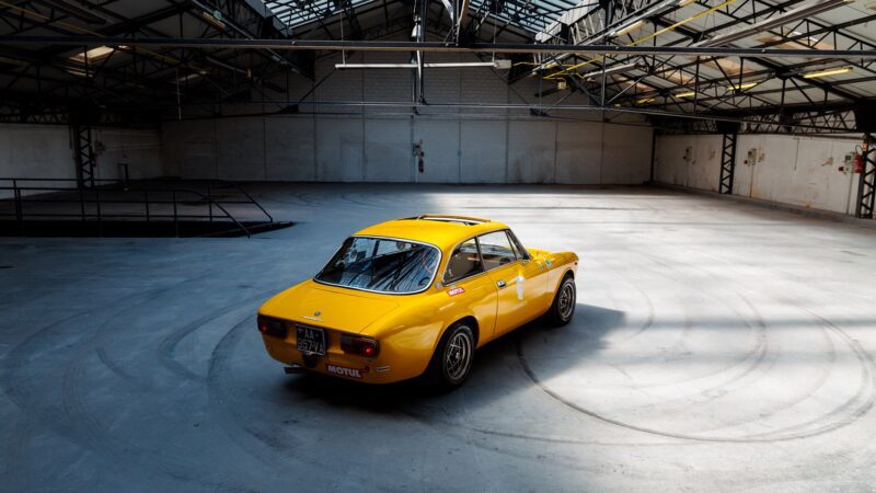 A yellow Alfa Romeo Giulia GTV vintage sports car is parked in the center of an empty industrial warehouse with large windows and a high ceiling.