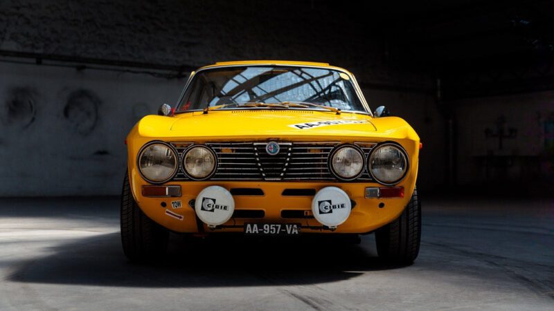 Front view of a yellow vintage Alfa Romeo Giulia GTV parked indoors with rally lights and a license plate reading "AA-957-VA"—a true Petrolicious moment for classic car enthusiasts.