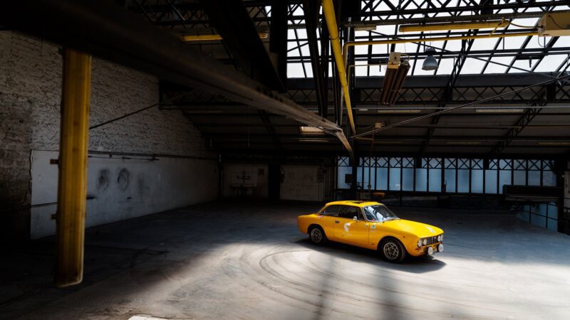 A yellow Alfa Romeo Giulia GTV is parked in the center of an empty industrial warehouse, illuminated by sunlight streaming through the roof windows—a scene worthy of Petrolicious.