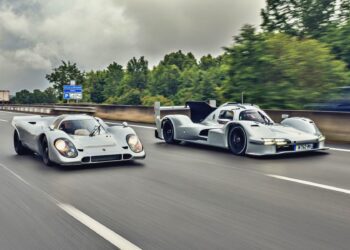 Two white Porsche 963 race cars, part of a Penske Tribute, drive side by side on a highway with trees and road signs in the background.
