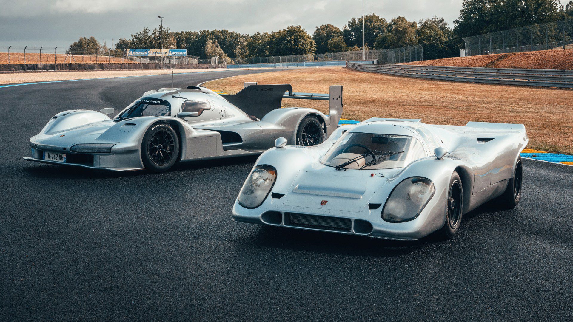 Two silver Hypercars are parked on a racetrack, with grass and trees in the background under a cloudy sky, capturing the spirit of FIA WEC and Le Mans racing.