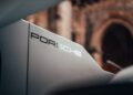 Close-up of a car spoiler with "Porsche" printed on it, set against a blurred old stone building—highlighting the unique 1-of-1 Porsche 963 RSP that breaks cover as a tribute to Penske ahead of Le Mans 2025.
