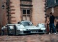 A silver 1-of-1 Porsche 963 RSP, crafted as a tribute to Penske ahead of Le Mans 2025, is parked on a cobblestone street with a man in a black shirt nearby and several people in the background.