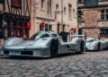 Two silver race cars, including the 1-of-1 Porsche 963 RSP Breaks Cover As A Tribute To Penske Ahead Of Le Mans 2025, are parked on a cobblestone street in a historic European town with old stone and timber buildings in the background.
