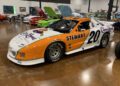 Orange and white IROC All-Star race car with the number 20 and "Stewart" on the side, parked indoors among other classic cars, reminiscent of legends raced at Laguna Seca during events like the Rolex Monterey Motorsports Reunion.