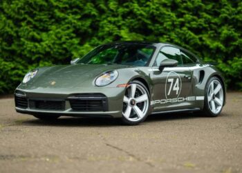 A gray Porsche 911 Turbo sports car with "74" and "Porsche" decals is parked on a paved surface in front of a dense green hedge.
