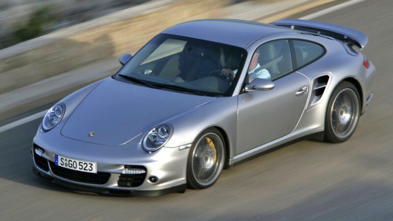 A silver Porsche 911 Turbo, viewed from above and slightly to the side, speeds down the road—The Last Porsche 911 Turbo To Feature A Manual Transmission.