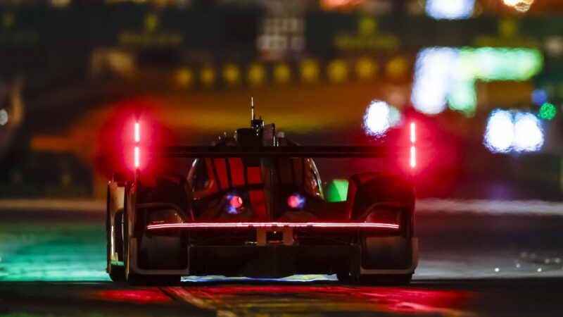 A race car with illuminated rear lights drives on a track at night, with blurred lights in the background, as Ferrari clinches a historic third straight Le Mans victory.