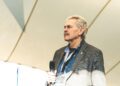 An older man in a blazer holding a microphone stands under a tent-like structure at the Festival of Speed, looking ahead with a serious expression.