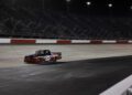 A black and red Ram race truck with the number 25, powered by HEMI V8s, drives alone on a lit NASCAR racetrack at night, with empty grandstands in the background.