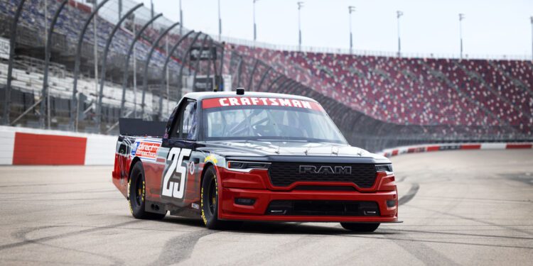 A black and red Ram race truck with the number 25 on the side, powered by HEMI V8s, drives on a racetrack with empty grandstands in the background.