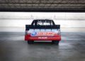 A rear view of a red and white Ram NASCAR race truck with "Direct Connection" branding, featuring powerful HEMI V8s, parked inside a garage facing a racetrack.