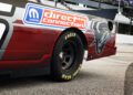 Close-up of a race car's rear side, featuring a Goodyear Eagle tire, "Direct Connection" and Mopar logos, part of a stylized graphic, and hints of its powerful HEMI V8 engine, parked on a racetrack.