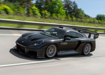 A matte black Porsche sports car with a large rear wing drives on a highway, surrounded by greenery and trees—a scene reminiscent of an RML Group P39 Hypercar or a Graham Rahal Performance masterpiece from a premier Hypercar Dealer.