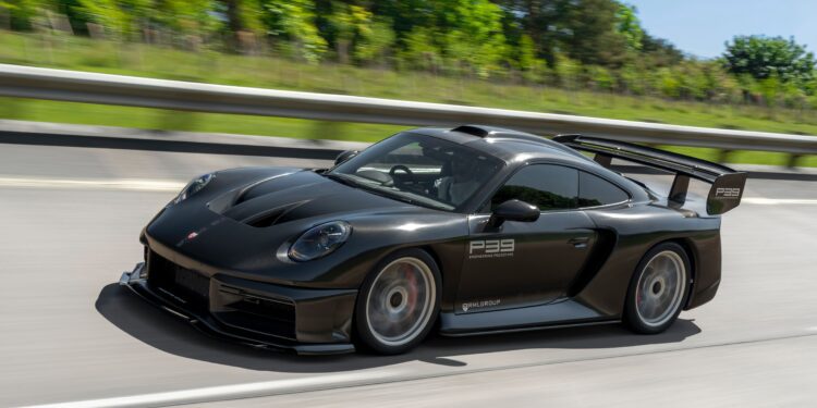 A matte black Porsche sports car with a large rear wing drives on a highway, surrounded by greenery and trees—a scene reminiscent of an RML Group P39 Hypercar or a Graham Rahal Performance masterpiece from a premier Hypercar Dealer.