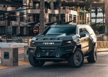 A matte black Rezvani SUV with angular, armored design—a unique creation—rests on a modern urban street in front of contemporary buildings and palm trees.