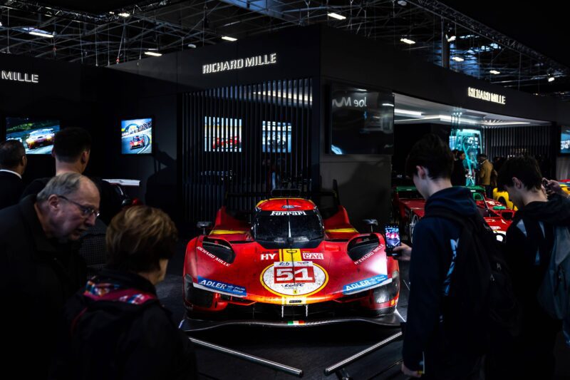 A red Ferrari racing car with number 51 is displayed at an indoor auto show in Manhattan, surrounded by visitors taking photos and admiring the European elegance of renowned exhibits.