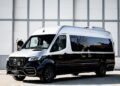 A black and white Mercedes-Benz Sprinter van, a unique creation, is parked outside in front of a large, gray industrial building that resembles a garage.